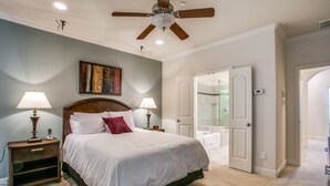 Een slaapkamer met een bed, twee bedlampen, een plafondventilator, een schilderij aan de muur, en een badkamer zichtbaar door een open deur.