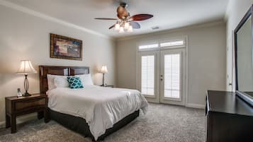 A bedroom with a large bed, two bedside tables with lamps, a ceiling fan, a painting on the wall, and a door with blinds.