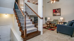 A spacious living area with a wooden staircase, a ceiling fan, and a modern abstract painting on the wall. - Beautifully furnished TownHome at shops (Plano)
