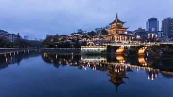 A traditional building with a multi-tiered roof, situated by a calm body of water reflecting lights, with modern buildings in the background.