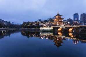 A traditional building with a multi-tiered roof, situated by a calm body of water reflecting lights, with modern buildings in the background.