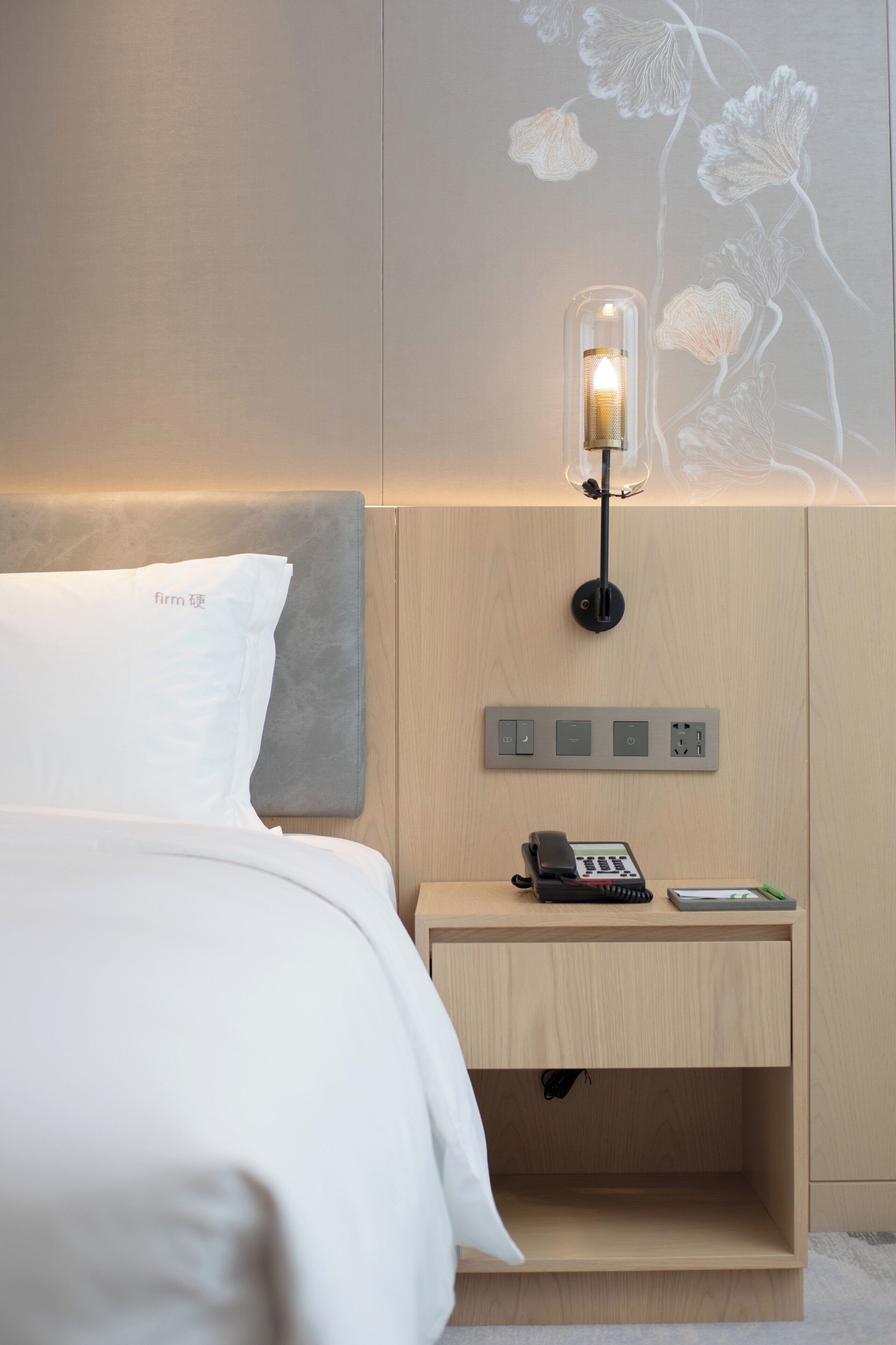A modern hotel room with a neatly made bed, a bedside table with a phone and a remote control, and a wall-mounted light fixture.