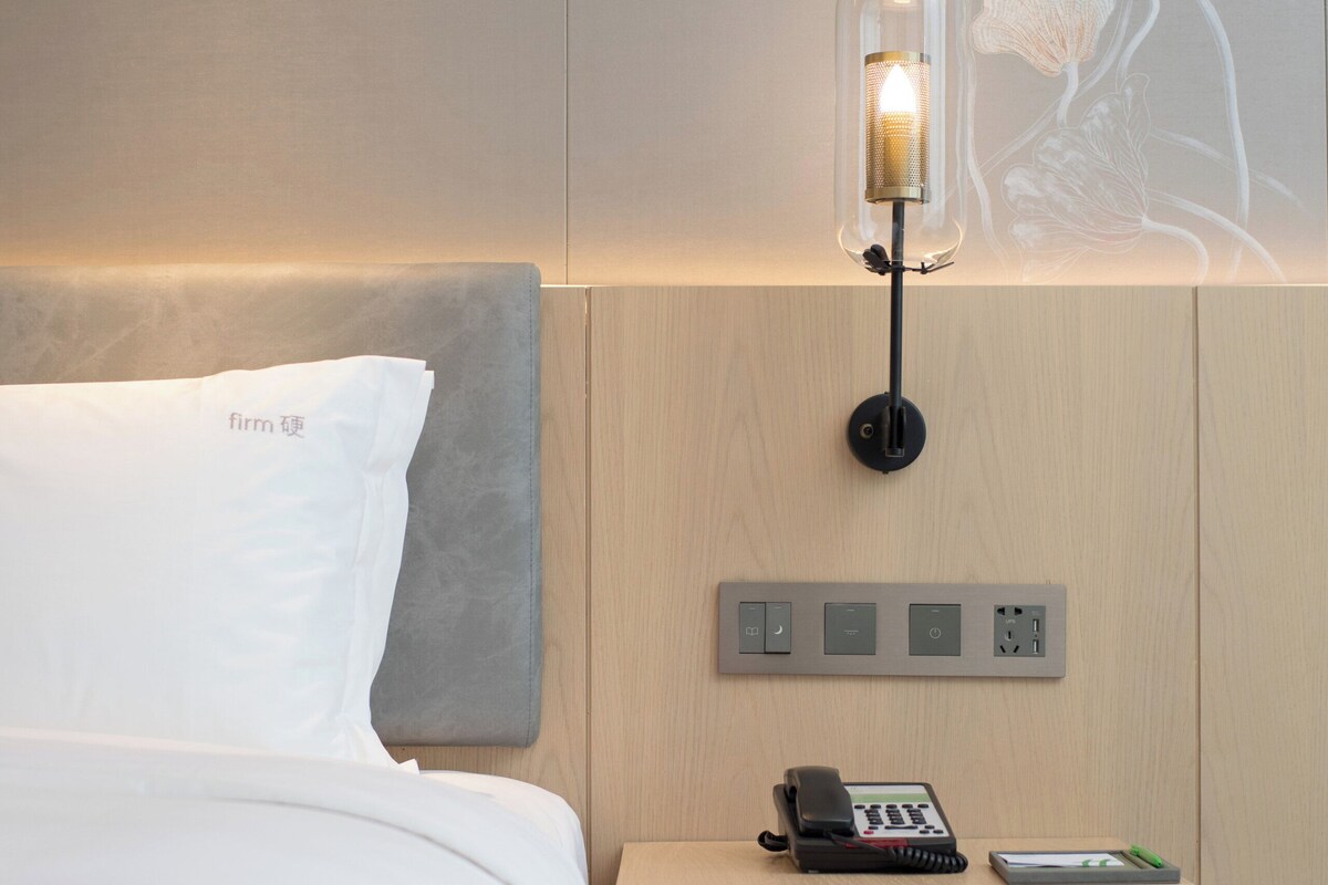 A modern hotel room with a neatly made bed, a bedside table with a phone and a remote control, and a wall-mounted light fixture.