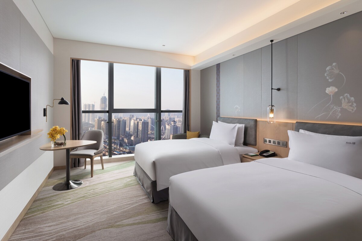 A modern hotel room with two beds, a desk, a chair, and a large window offering a city view.