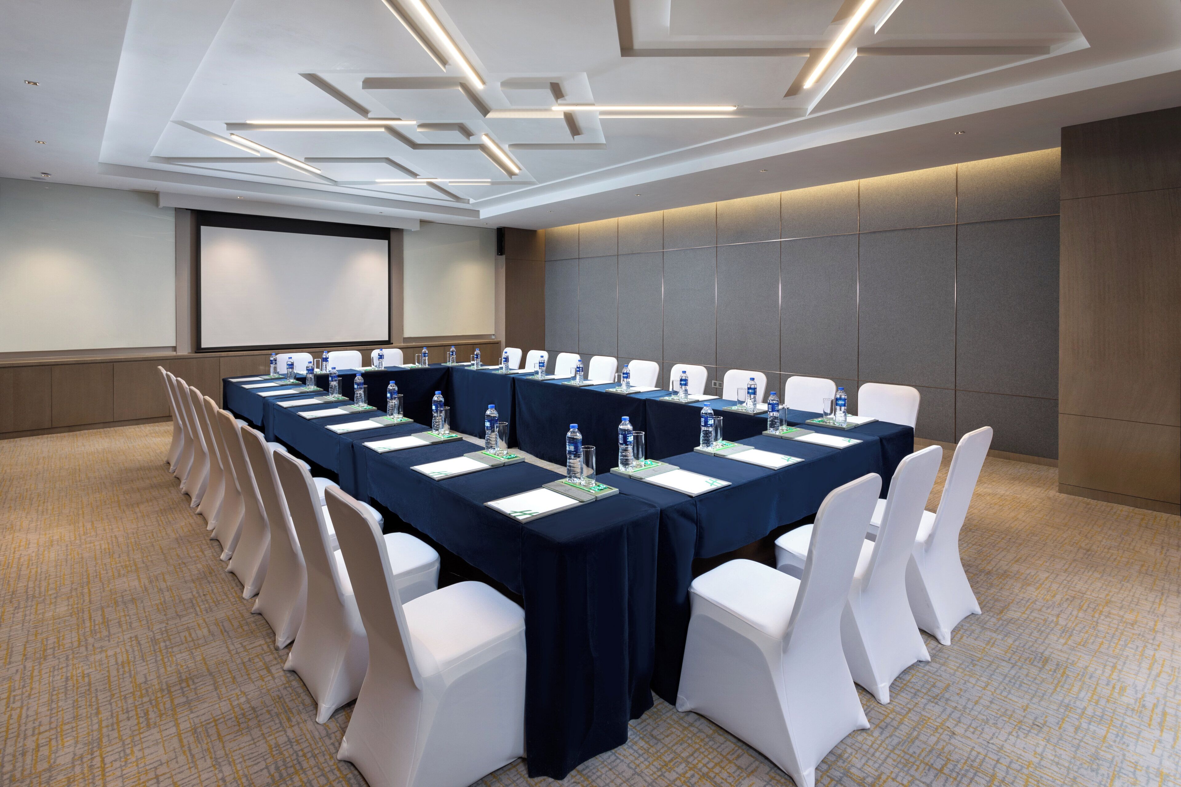 A conference room with a long table, white chairs, and a projector screen.