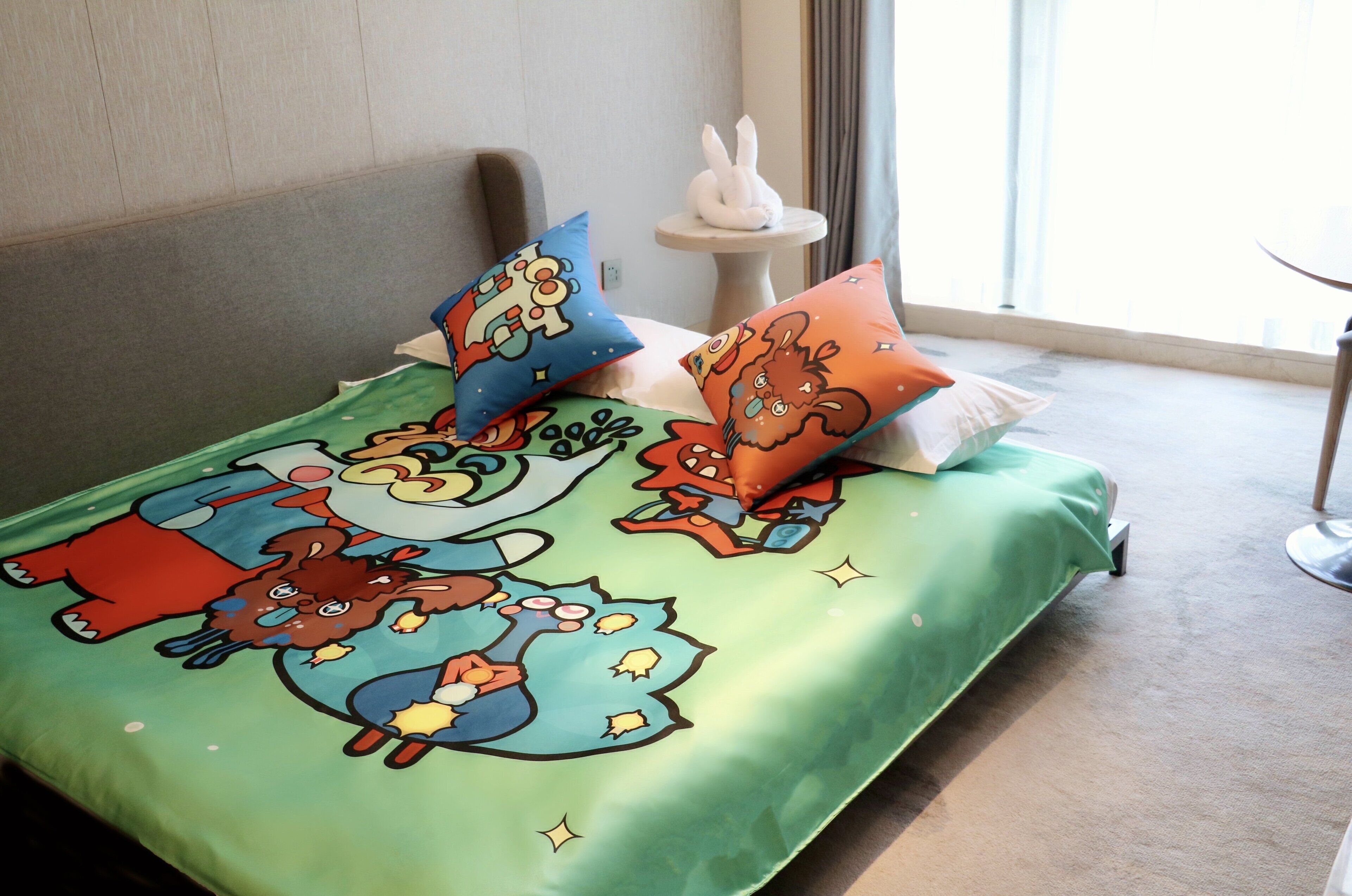 Una cama con una colcha verde adornada con personajes de dibujos animados, un almohadón blanco y una mesita redonda con una escultura de conejo.