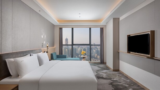 Ein modernes Hotelzimmer mit einem großen Bett, einem an der Wand montierten Fernseher und Blick auf die Stadt durch das Fenster.