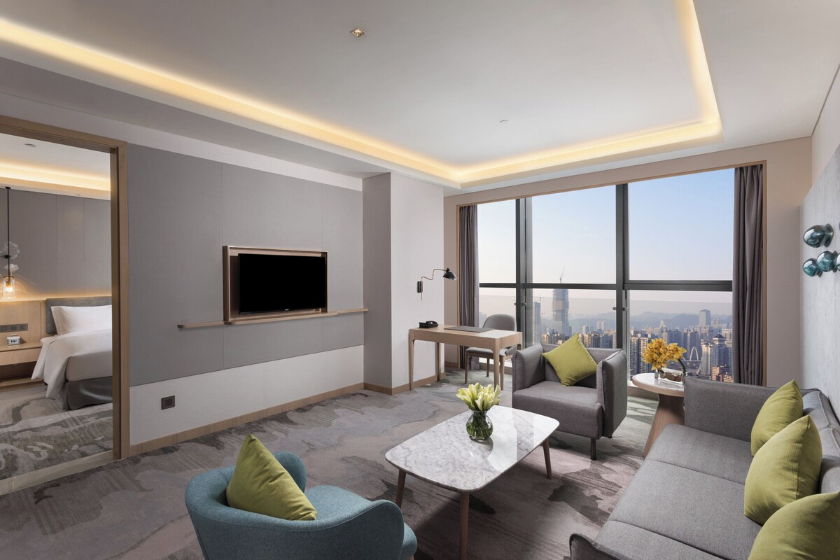 A modern hotel room with a large window, a flat-screen TV, a sofa, a coffee table, and a desk.