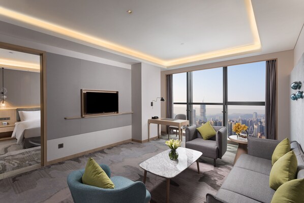 A modern hotel room with a large window, a flat-screen TV, a sofa, a coffee table, and a desk.