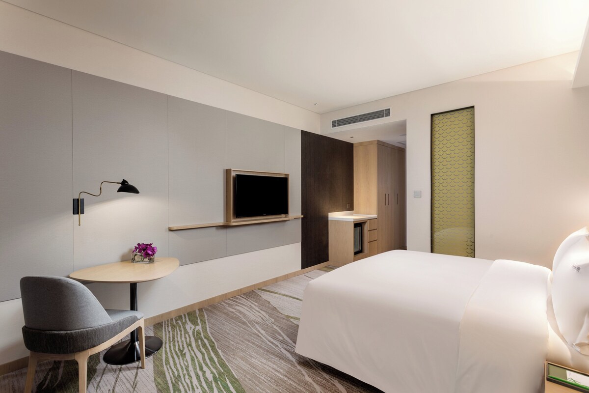 A modern hotel room with a bed, a desk, a chair, a television, and a small table with a vase of flowers.