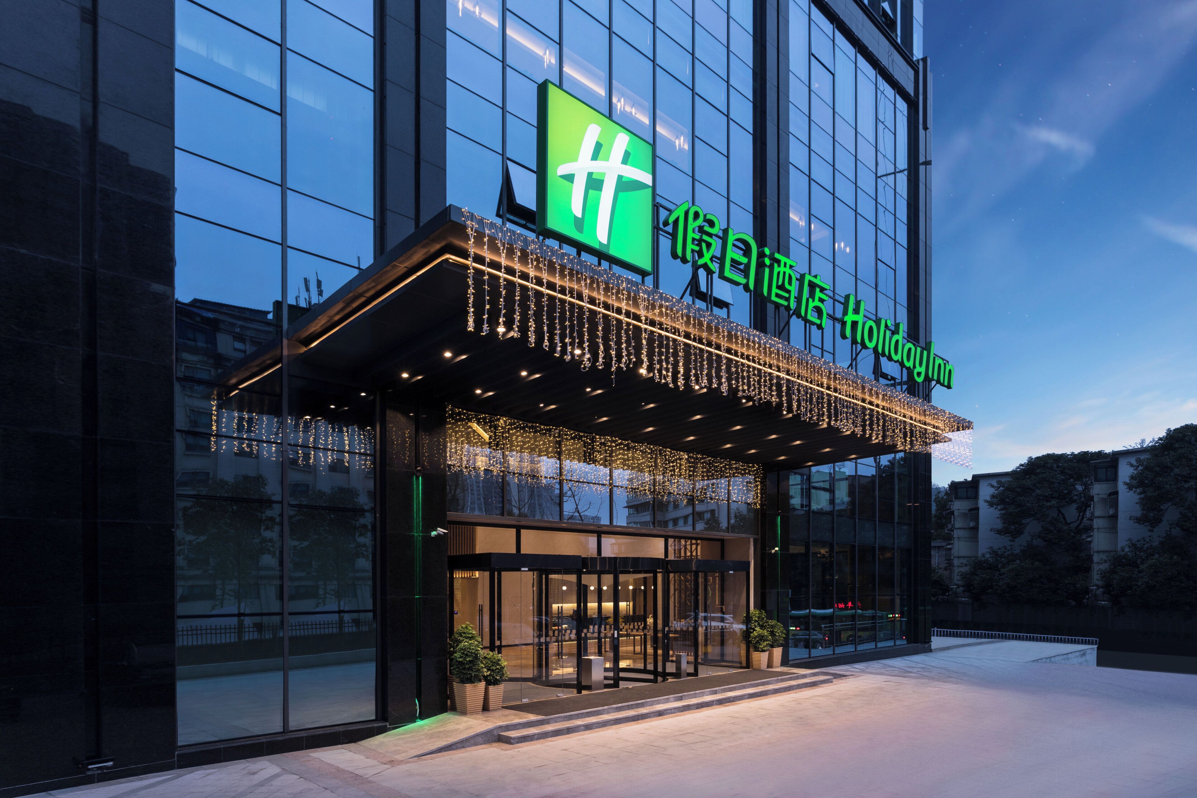 A hotel entrance with a glass facade, a green sign with white text, and a covered entrance area with lights.