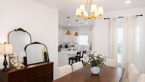 Un espace repas moderne avec une table en bois, des chaises blanches, un vase de fleurs et un grand miroir placé sur un buffet en bois foncé.