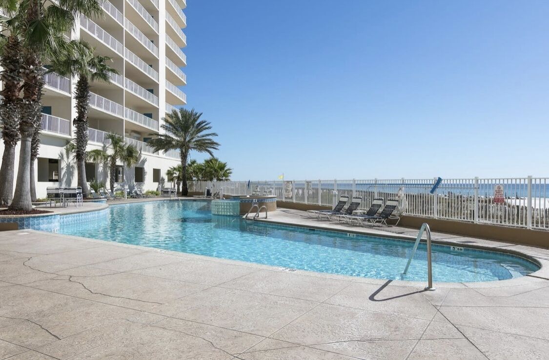 Une piscine d’hôtel offrant une vue dégagée sur l’océan, entourée de palmiers et d’une clôture blanche.