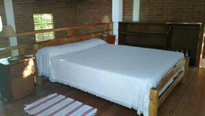 A bedroom with a wooden bed, a nightstand, a lamp, and a brick wall. - Eluney Cabanas (Mina Clavero)