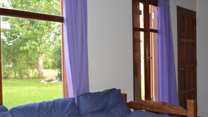 A room with a wooden bed frame, a blue mattress, and a view of a green outdoor area through a window with purple curtains. - Eluney Cabanas (Mina Clavero)