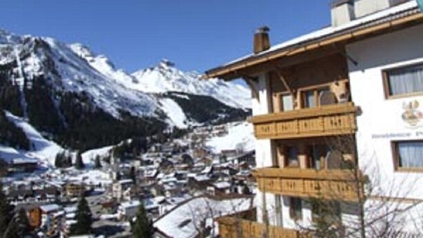 Un pueblo de montaña con picos nevados al fondo y un edificio con balcones en primer plano.