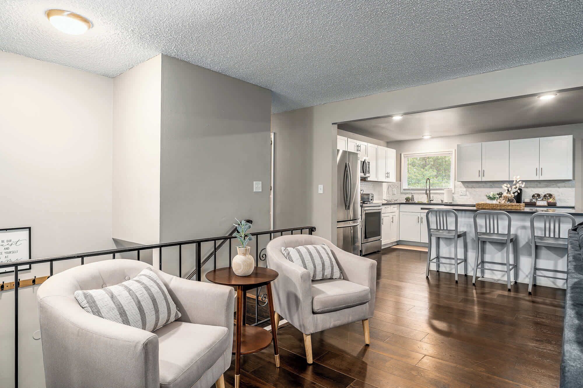 A modern kitchen with stainless steel appliances, a bar area with bar stools, and a staircase leading to a living area with two armchairs and a small round table.