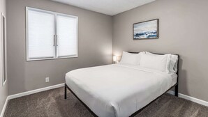 A bedroom with a bed, a window with blinds, a wall-mounted painting, and a ceiling light.