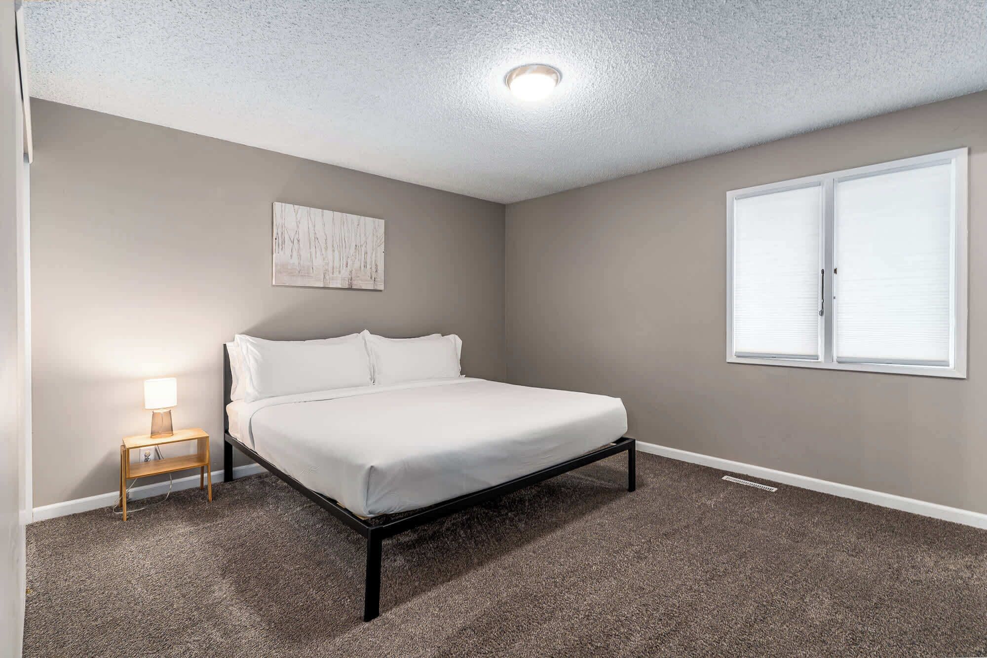 A bedroom with a bed, a nightstand, a wall-mounted artwork, a window with blinds, and a ceiling light.