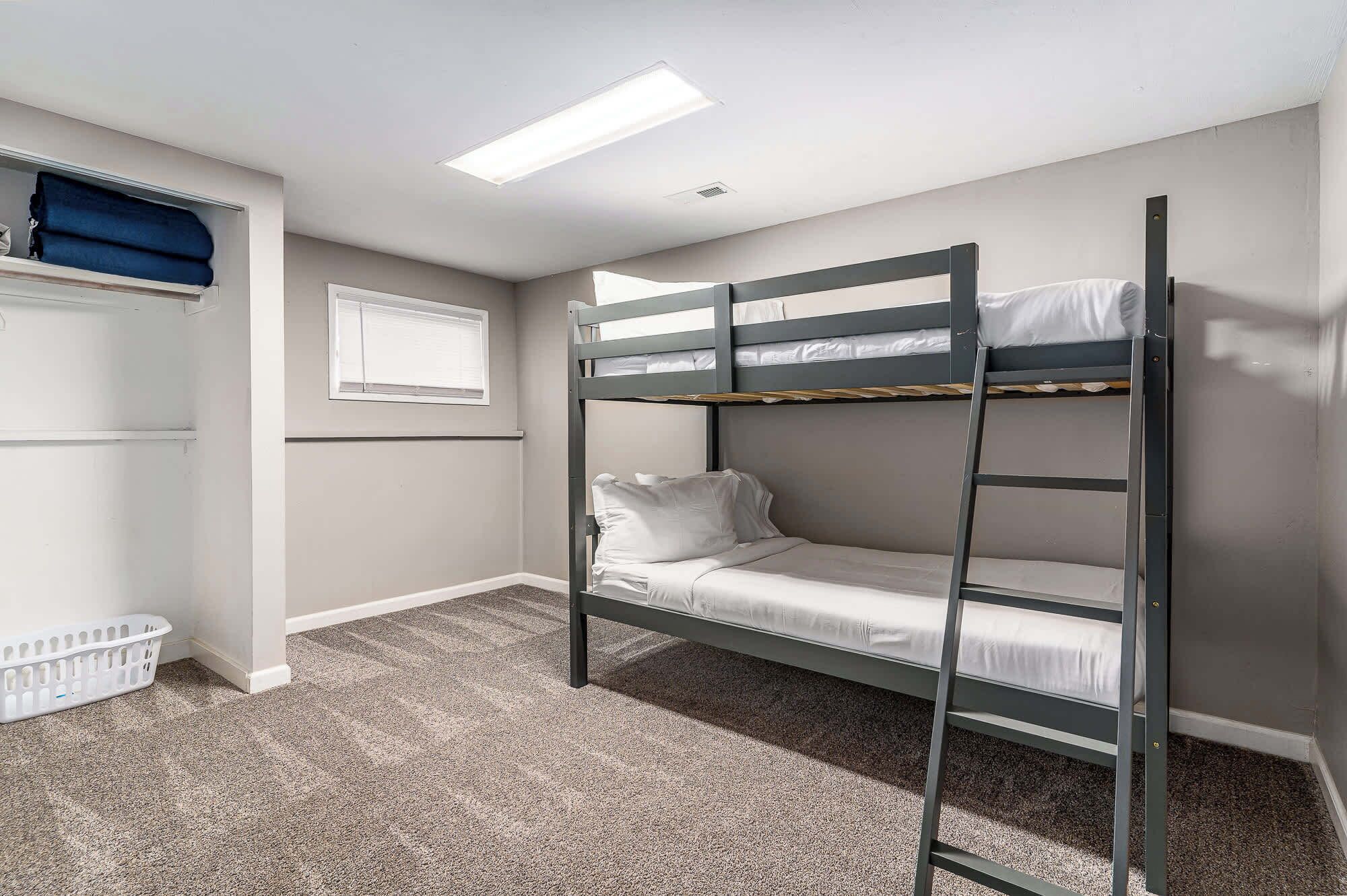 A bunk bed room with a carpeted floor, a window with blinds, and a laundry basket.