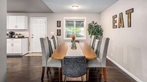 A dining area with a wooden table and grey upholstered chairs, a kitchen with white cabinets, and a window with a view of greenery.