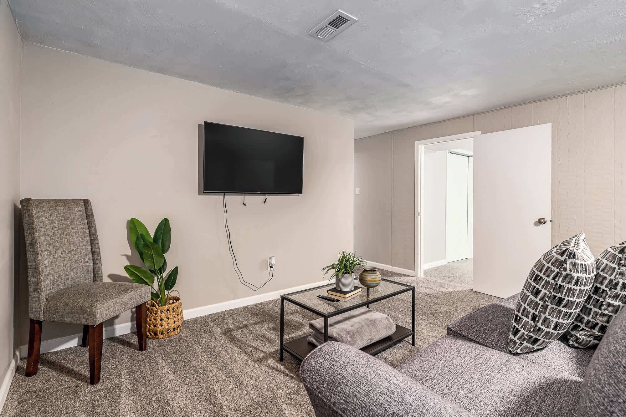 A living room with a flat-screen TV mounted on the wall, a sofa, a chair, a coffee table with a plant, and a door leading to another room.