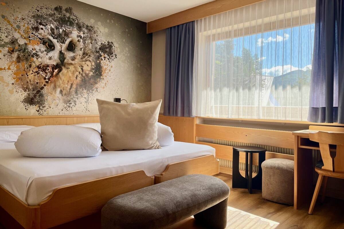 A modern hotel room with a large bed, a small table, and a chair. There is a window with curtains and a view of the outdoors.