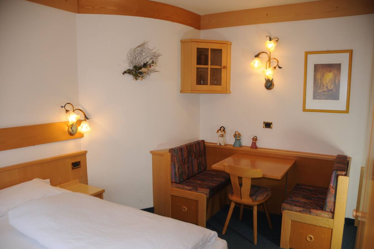 A small, cozy room with a bed, a wooden bench with a cushioned seat, a small table, and a cabinet.