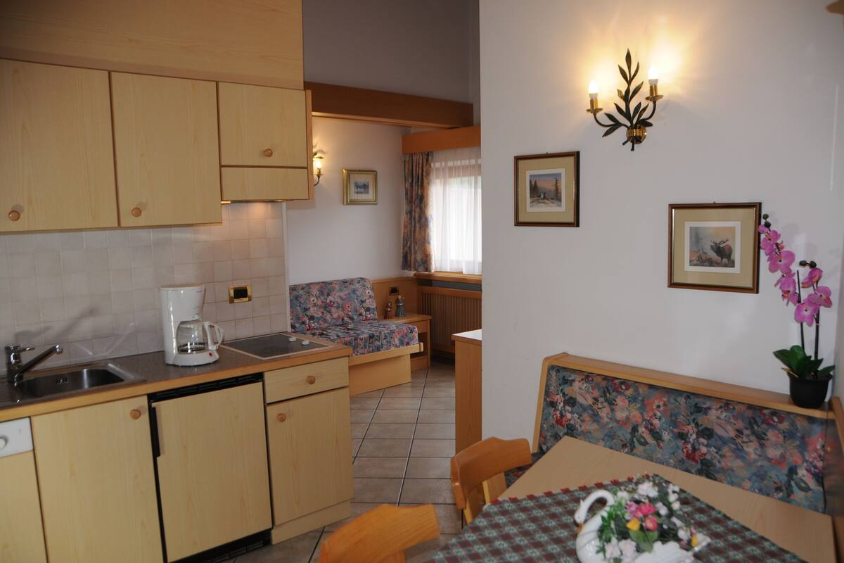 A compact kitchen with wooden cabinets, a coffee maker, and a dining area with a floral-patterned table and bench.