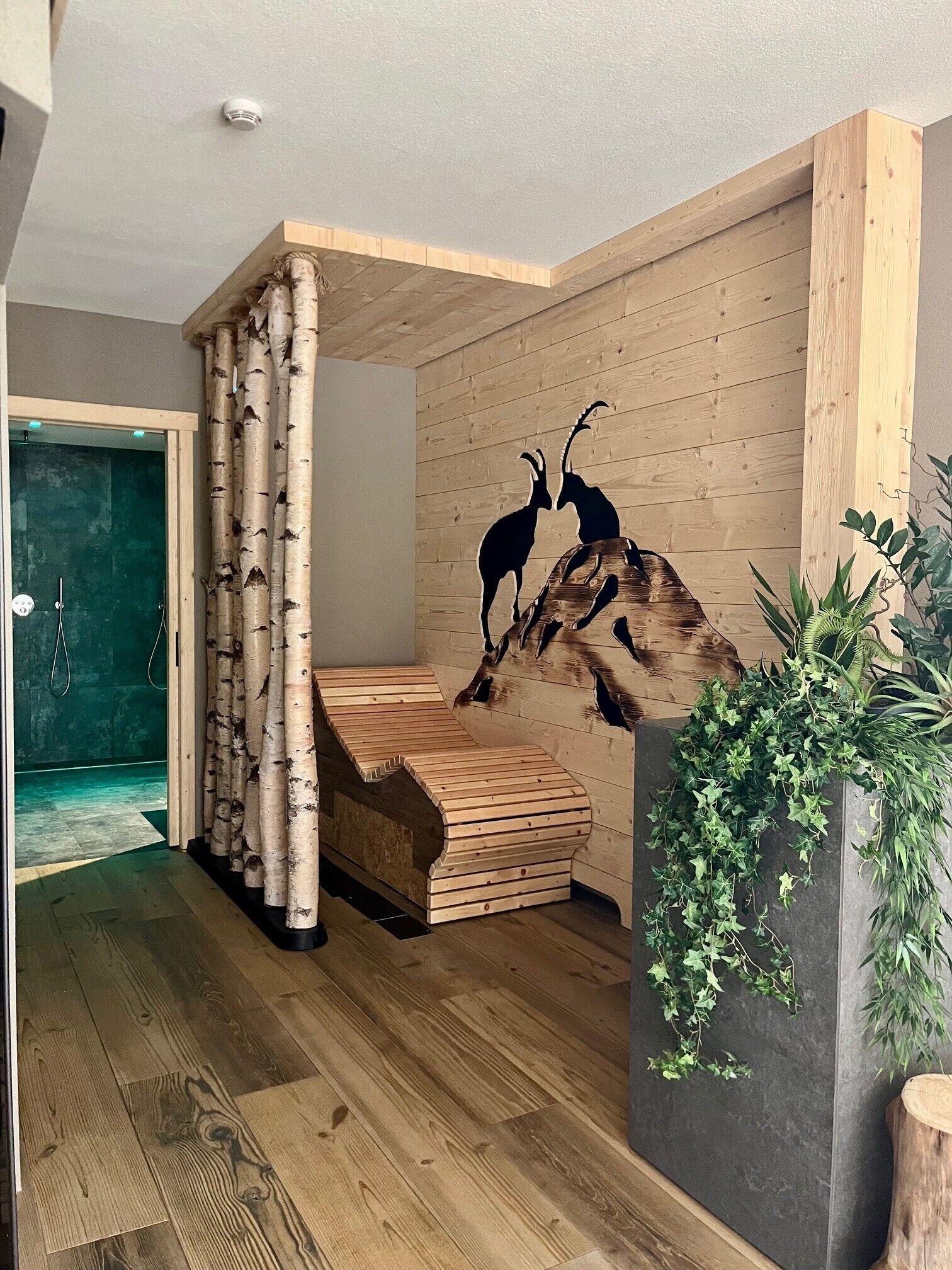 Une pièce moderne avec un plancher en bois, un banc en bois, des rideaux ornés de motifs de bouleaux, une sculpture d’antilopes fixée au mur et une plante en pot.