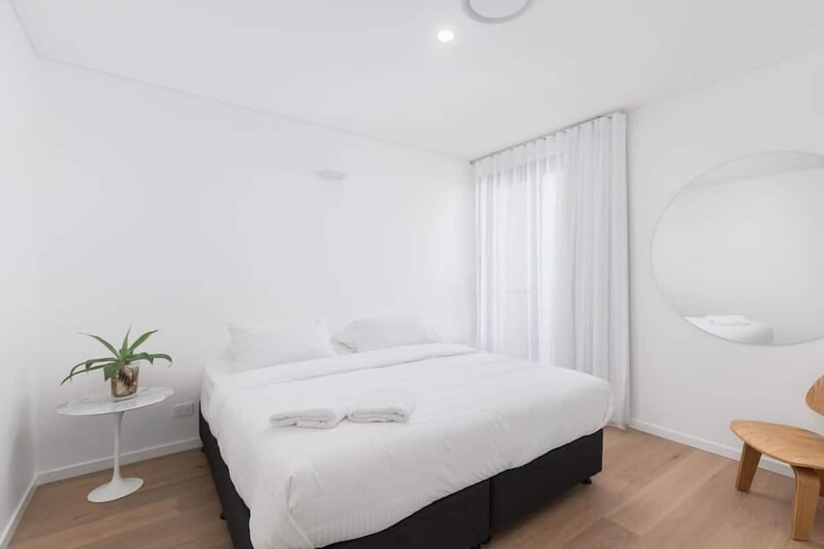 Ein modernes Hotelzimmer mit einem Bett, einem kleinen runden Tisch mit Pflanze, einem Sessel und einem runden Spiegel an der Wand.