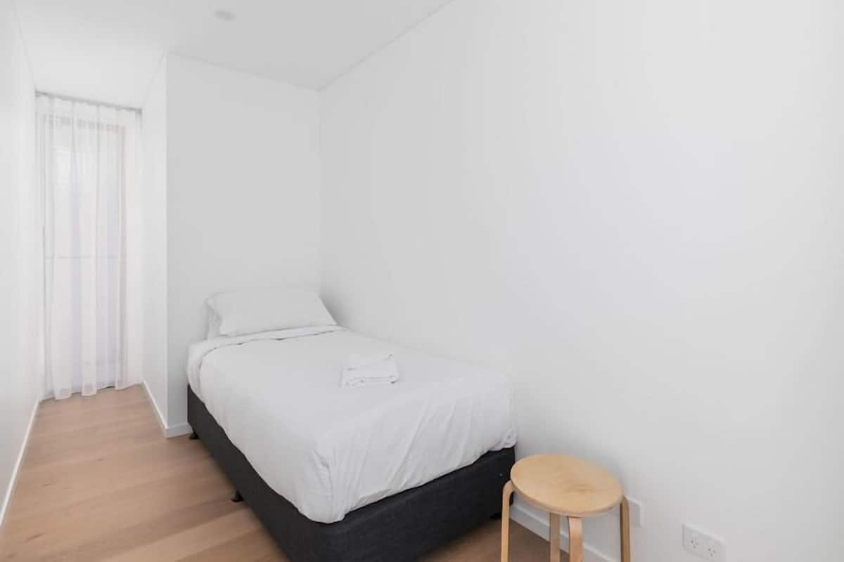 Ein schlicht eingerichtetes Schlafzimmer mit einem Bett, einem kleinen Hocker und einem Fenster mit transparenten Vorhängen.