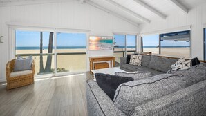 Ein geräumiges Wohnzimmer mit großem Fenster zum Strandblick, ein Korbstuhl, ein graues Sofa und ein Holztisch.