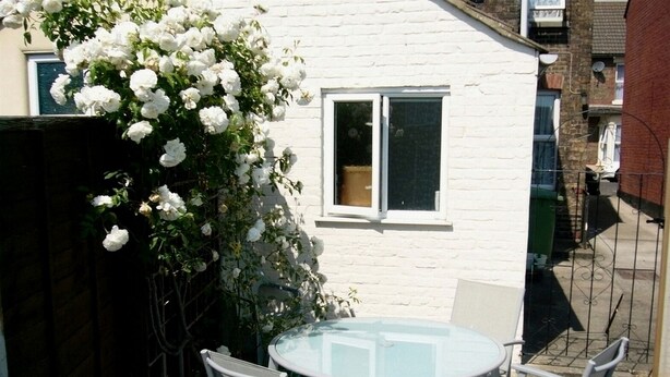 A round glass table with four chairs, a white wall, and a white flowering bush.
