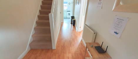 A hallway with a wooden floor, a staircase, and a radiator. There are signs on the wall and a small table with a laptop.
