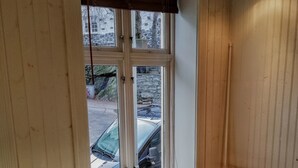 Ein Zimmer mit einem weißen Tisch und zwei Holzsesseln, ein Fenster mit Blick auf ein parkendes Auto und eine Mauer aus Stein.