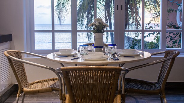 Un coin repas avec une table ronde, quatre chaises en osier, un vase rempli de fleurs et une vue sur la mer grâce à de grandes fenêtres.