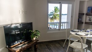Un salon avec un téléviseur, une plante, une table à manger avec des chaises et une vue sur la mer par une fenêtre.