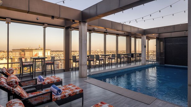 An indoor swimming pool area with lounge chairs and tables, large windows offering a city view, and a modern architectural design. - Courtyard by Marriott Amritsar (Amritsar)