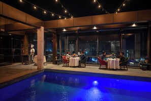 Un espace repas aménagé au bord de la piscine, avec des tables et des chaises, une piscine et de grandes fenêtres offrant une vue sur la ville.