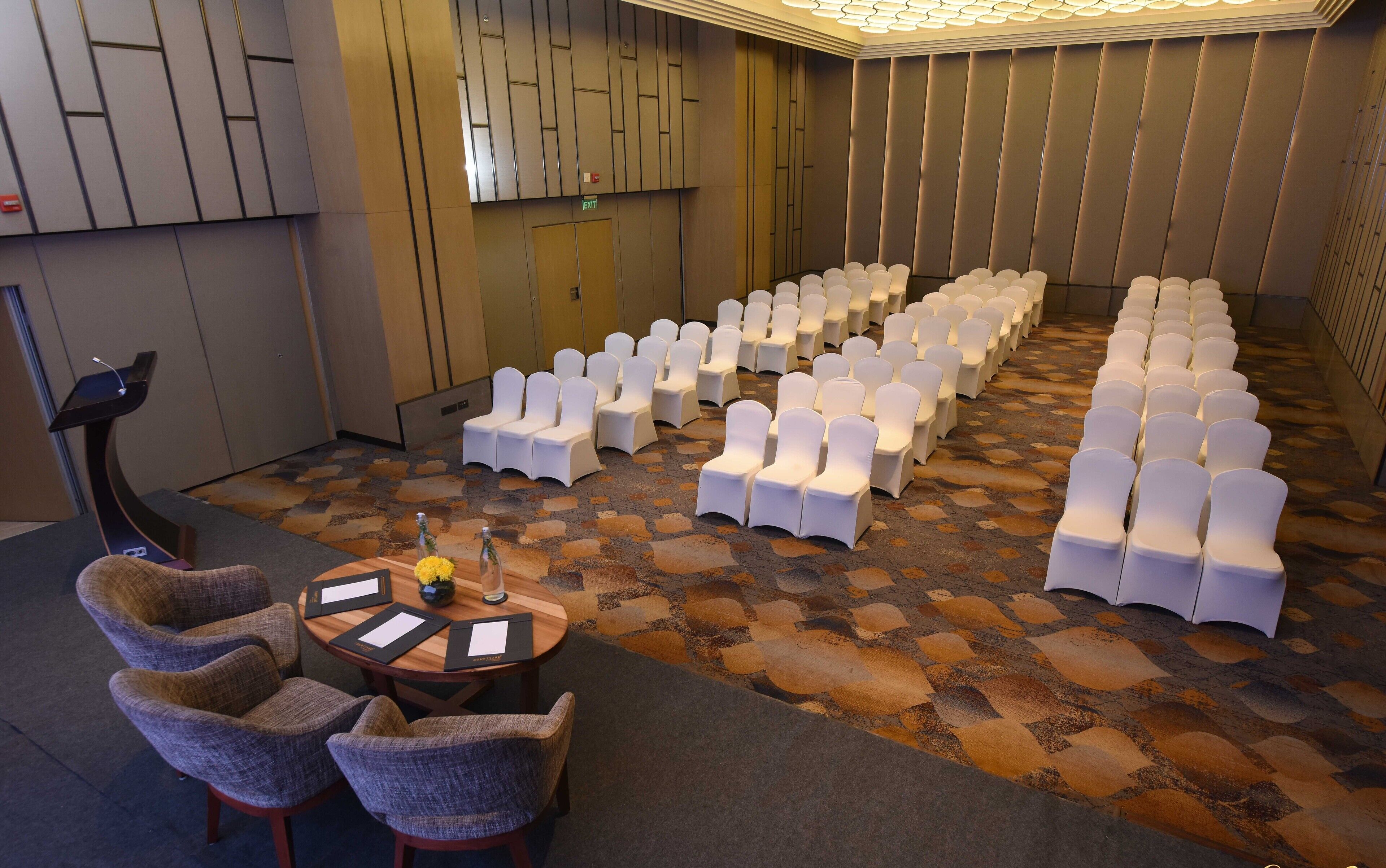 A conference room with rows of white chairs, a small table with documents, and a speaker.