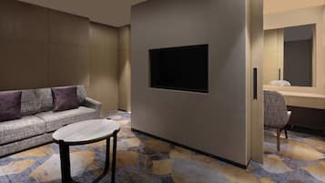 A modern hotel room with a sofa, a small round table, a flat-screen TV mounted on the wall, and a desk with a chair.