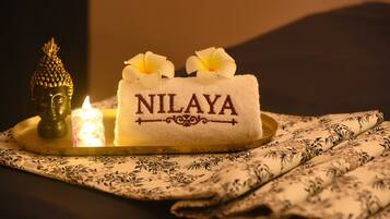 Una toalla con el bordado "NILAYA", una estatua de Buda, una vela encendida y un textil estampado.