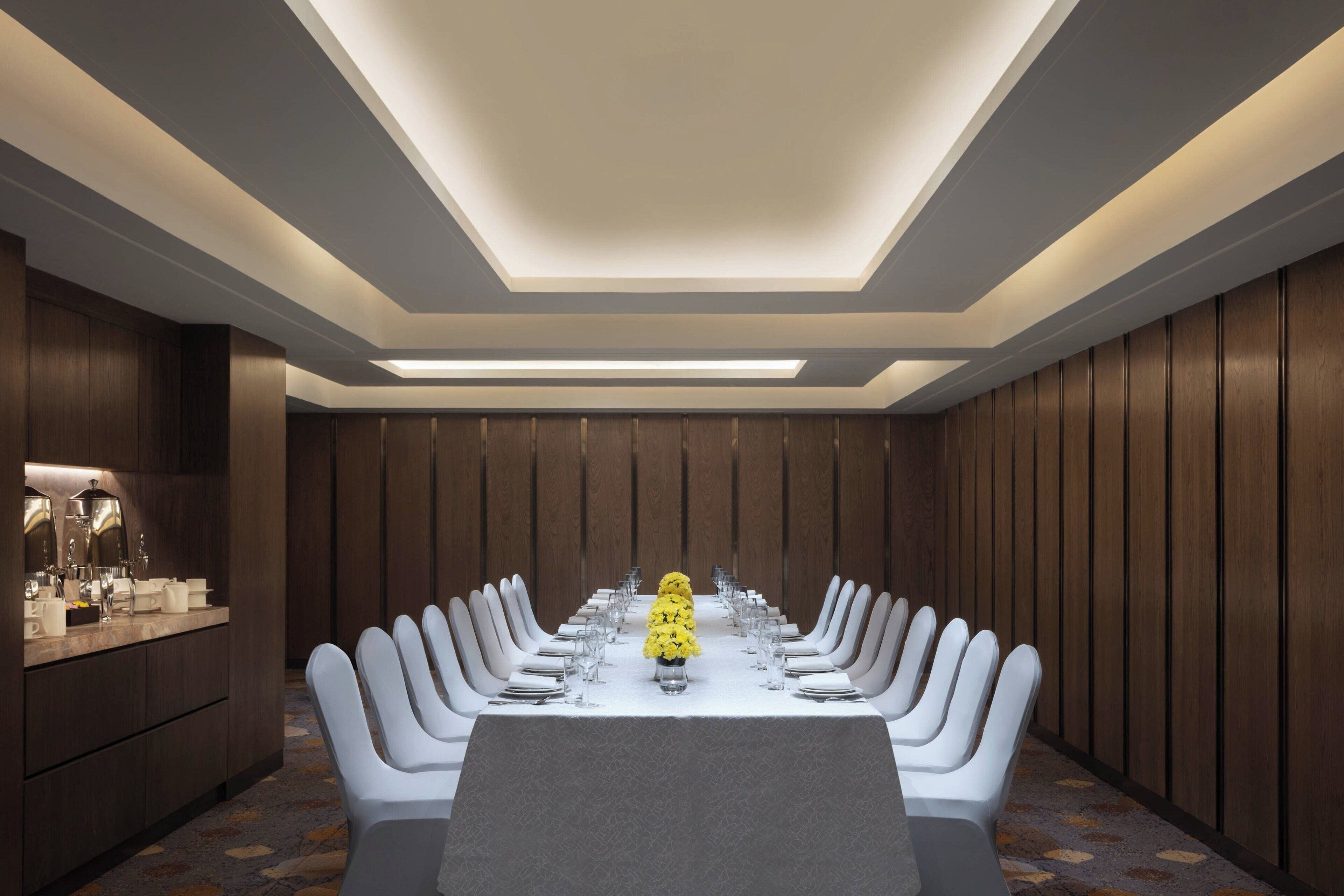 Un salón de conferencias con una larga mesa, sillas blancas y un arreglo floral amarillo en el centro.
