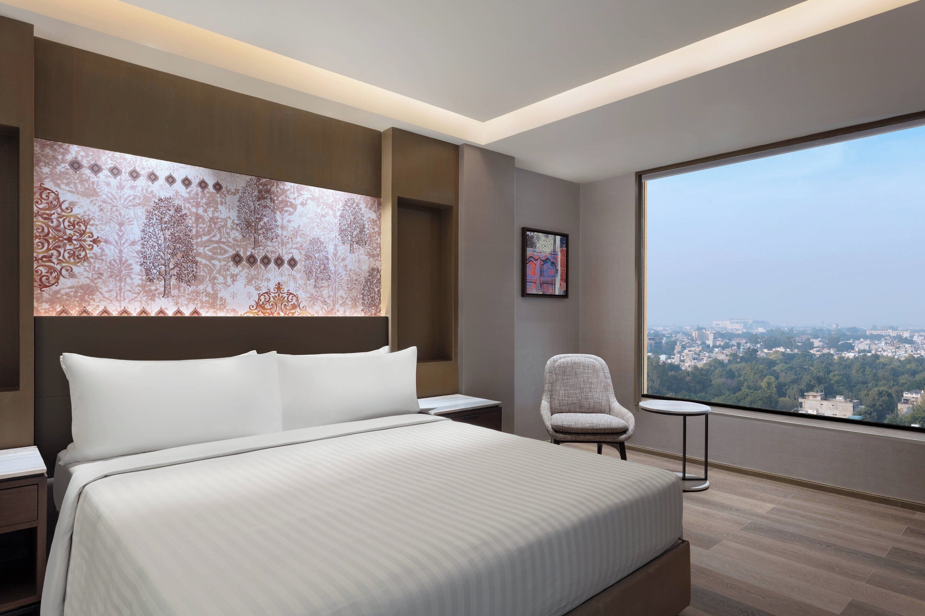 Habitación de hotel moderna con una cama grande, una silla, una mesita y un ventanal con vistas a la ciudad.