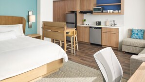 Quarto de hotel moderno com cama, área de refeições com mesa e cadeiras, cozinha compacta e sala de estar com sofá.