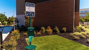A sign instructing to clean up after dogs, a green trash bin, and a well-maintained grassy area with shrubs and a building in the background.