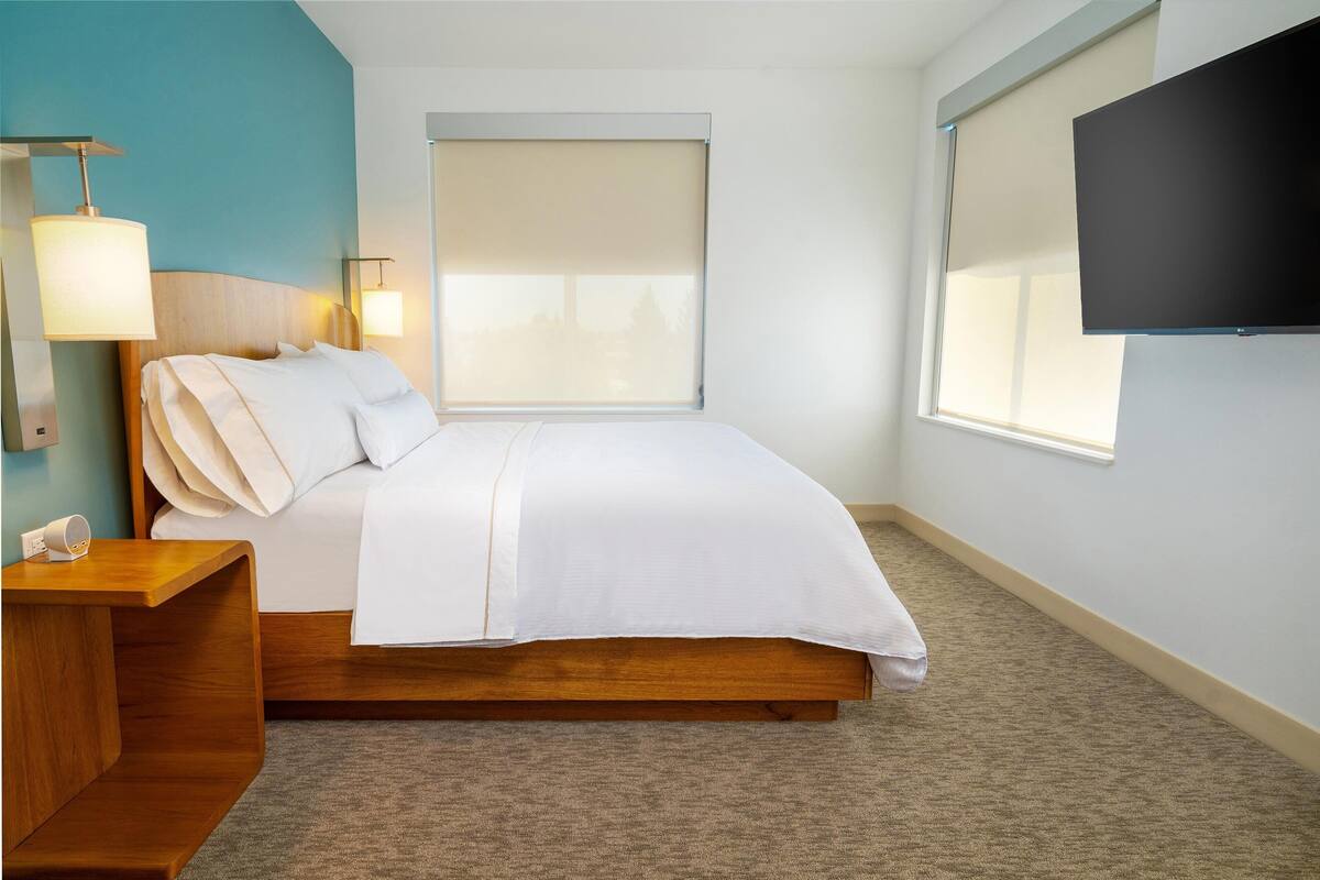 Una cama bien hecha en una habitación de hotel con cabecera de madera, una mesita de noche con lámpara, un ventanal con vistas y un televisor de pantalla plana montado en la pared.
