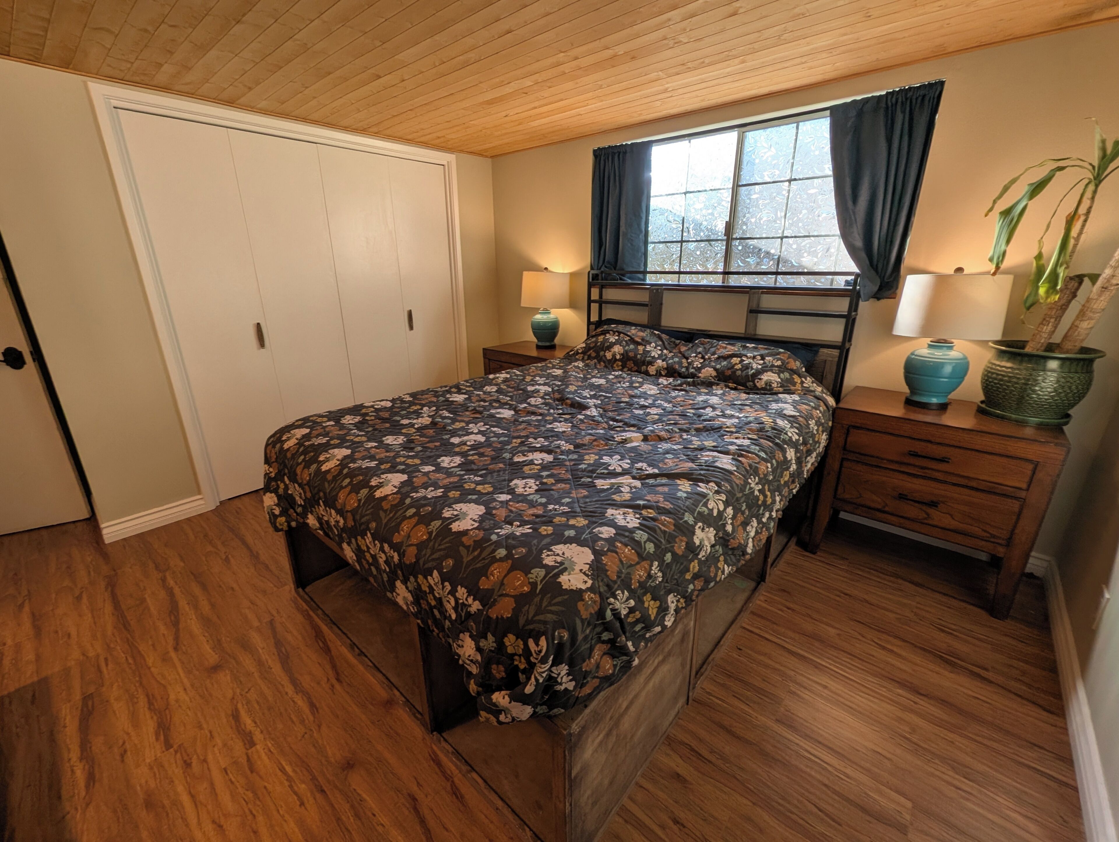 A bedroom with a wooden floor, a bed with a floral bedspread, a nightstand with a lamp, a dresser, and a window with curtains.