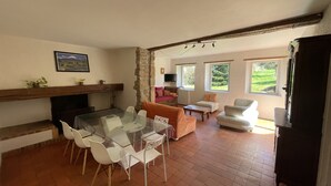 Un vaste espace de vie comprenant une table à manger en verre, des chaises blanches, une cheminée et un mur en pierre. Deux fenêtres laissent entrer la lumière naturelle et offrent une vue sur la verdure environnante.